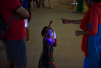 A child holds a glowing light toy while an adult points at it. The setting appears to be a dimly lit outdoor area, possibly during an evening event or festival. The child is wearing a colorful dress with floral patterns, and the adults nearby are dressed casually.