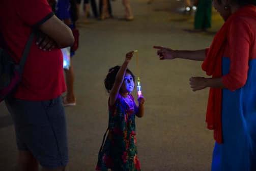 A child holds a glowing light toy while an adult points at it. The setting appears to be a dimly lit outdoor area, possibly during an evening event or festival. The child is wearing a colorful dress with floral patterns, and the adults nearby are dressed casually.
