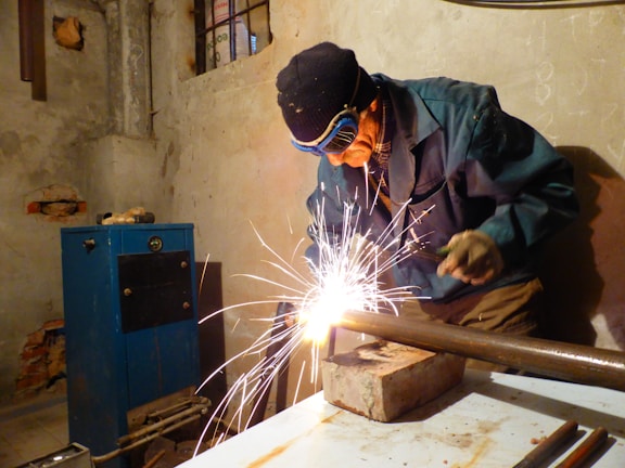 A person wearing protective goggles and gloves is engaged in welding a metal pipe on a workbench, surrounded by sparks. The scene takes place in a dimly lit workshop with raw walls and visible utility equipment.