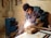 A person wearing protective goggles and gloves is engaged in welding a metal pipe on a workbench, surrounded by sparks. The scene takes place in a dimly lit workshop with raw walls and visible utility equipment.
