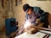 A person wearing protective goggles and gloves is engaged in welding a metal pipe on a workbench, surrounded by sparks. The scene takes place in a dimly lit workshop with raw walls and visible utility equipment.