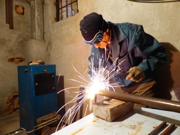 A person wearing protective goggles and gloves is engaged in welding a metal pipe on a workbench, surrounded by sparks. The scene takes place in a dimly lit workshop with raw walls and visible utility equipment.