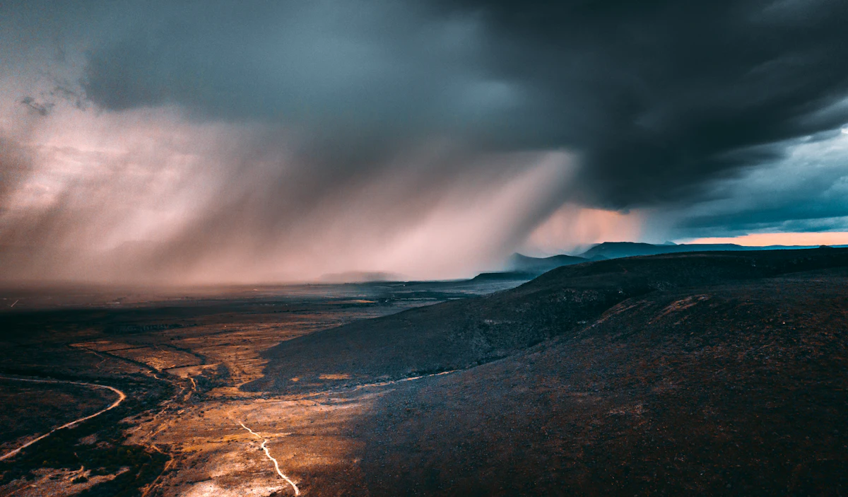 Rain falling from a dark storm cloud over the mountains and plains around Graaff-Reinet, with a road winding across the plain below