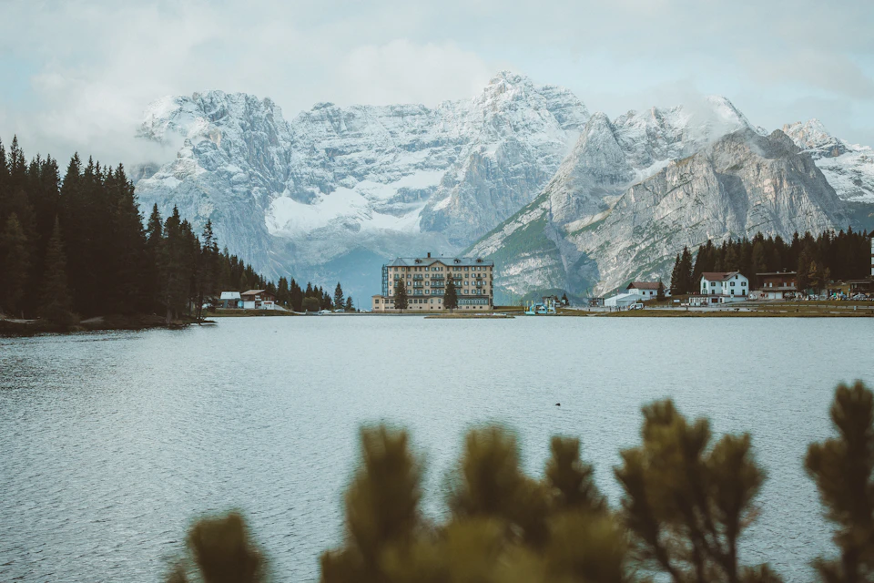 The image depicts a serene mountain landscape with a large lake at its center, surrounded by snow-capped mountains and lush green trees. In the distance, there is a large building nestled among the mountains, adding an element of human presence to the natural scene. The perspective of the photo is taken from across the lake, providing a panoramic view of the landscape.