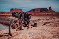 A rugged mountain bike is parked on rocky terrain with several packs attached to it, indicating it is prepared for a long journey. In the background, striking red rock formations rise against a clear blue sky, suggesting a desert landscape. Hiking boots are placed near the bike on the ground.
