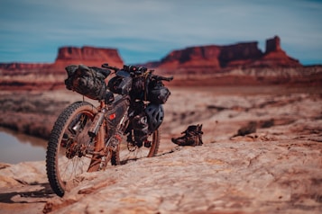 A rugged mountain bike is parked on rocky terrain with several packs attached to it, indicating it is prepared for a long journey. In the background, striking red rock formations rise against a clear blue sky, suggesting a desert landscape. Hiking boots are placed near the bike on the ground.