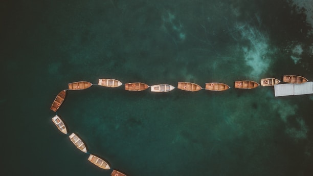 A series of small wooden boats are lined up on a dark, calm body of water, creating a gentle curve. The boats exhibit various shades of brown and are neatly tethered to a dock on the right side. The water displays a deep, murky green color with lighter patches, suggesting underlying textures or reflections.