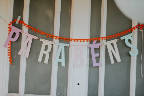 Colorful letters spell out 'PARABÉNS' strung together on a bright orange garland, displayed against a white paneled background.