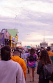 A crowded boardwalk filled with people walking in various directions. Colorful buildings, food stalls, and shops line the sides. The sky is overcast with a hint of sunlight breaking through the clouds, and a seagull is flying overhead.