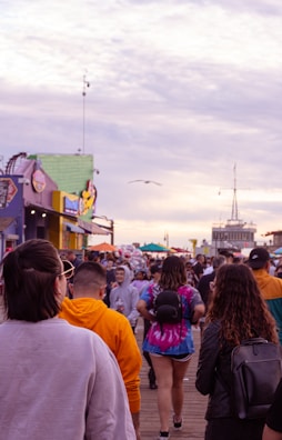 A crowded boardwalk filled with people walking in various directions. Colorful buildings, food stalls, and shops line the sides. The sky is overcast with a hint of sunlight breaking through the clouds, and a seagull is flying overhead.