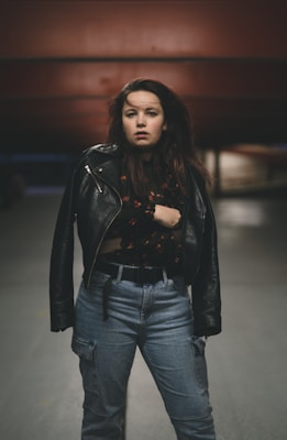 A person with long hair stands confidently, wearing a leather jacket over a patterned shirt and high-waisted jeans. The background appears to be an industrial or urban setting with muted tones.