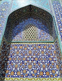 An intricate architectural detail showcases vibrant Persian tilework. The design features a rich combination of geometric patterns with floral motifs in blue, gold, and white. A pointed arch frames the decorative panel, marked by precise symmetry and elaborate ornamentation.