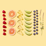 A bright and colorful arrangement of assorted fruits and herbs displayed on a light yellow background. The layout includes rows of raspberries, grapefruit slices, pineapple wedges, lime wedges, blueberries, and sprigs of lavender.