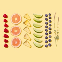 A bright and colorful arrangement of assorted fruits and herbs displayed on a light yellow background. The layout includes rows of raspberries, grapefruit slices, pineapple wedges, lime wedges, blueberries, and sprigs of lavender.