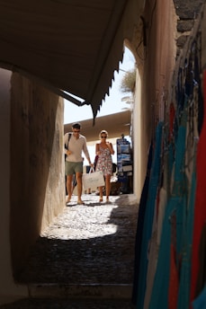 A man and a woman walk through a narrow, cobblestone alley lined with colorful garments on hangers. The man is wearing a white shirt and shorts, while the woman is in a floral dress, both carrying shopping bags. Sunlight casts shadows on the ground, enhancing the ambiance of a quaint shopping area.