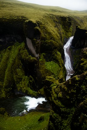A lush green landscape featuring a dramatic waterfall cascading down a steep, moss-covered cliff into a small pool. The surrounding terrain is rugged and covered in dense vegetation.