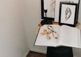 On a small round table, an open book is laid out with a wooden cross and a rosary placed on top. In the background, there are framed black and white artworks and a black candleholder. The setup is minimalistic with a serene ambiance.