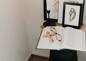 On a small round table, an open book is laid out with a wooden cross and a rosary placed on top. In the background, there are framed black and white artworks and a black candleholder. The setup is minimalistic with a serene ambiance.