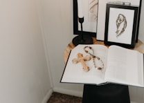 On a small round table, an open book is laid out with a wooden cross and a rosary placed on top. In the background, there are framed black and white artworks and a black candleholder. The setup is minimalistic with a serene ambiance.