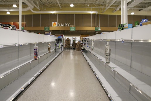The image depicts the interior of a grocery store with nearly empty shelves on both sides. The area is located in the dairy section of the store, as indicated by the overhead signage. There are a few remaining items scattered sparsely across the shelves. In the background, two individuals can be seen, possibly store employees, standing near a cart filled with boxes.