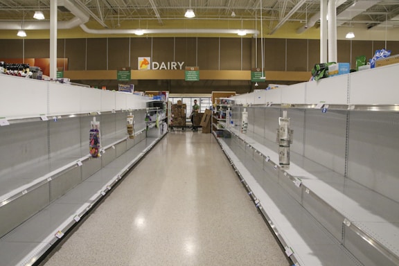 The image depicts the interior of a grocery store with nearly empty shelves on both sides. The area is located in the dairy section of the store, as indicated by the overhead signage. There are a few remaining items scattered sparsely across the shelves. In the background, two individuals can be seen, possibly store employees, standing near a cart filled with boxes.