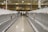 The image depicts the interior of a grocery store with nearly empty shelves on both sides. The area is located in the dairy section of the store, as indicated by the overhead signage. There are a few remaining items scattered sparsely across the shelves. In the background, two individuals can be seen, possibly store employees, standing near a cart filled with boxes.