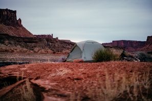 A camping scene set in a desert landscape features a small tent pitched on red rocky terrain. In the background, there are large rock formations and a wide, open sky. A bicycle is parked on the ground next to the tent, indicating a possible bike tour or adventure trip.