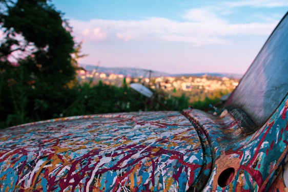 A paint-splattered, old car is parked in a natural setting, with a distant cityscape visible in the background. The vehicle's surface displays an abstract combination of colors against a backdrop of greenery and a blue sky.