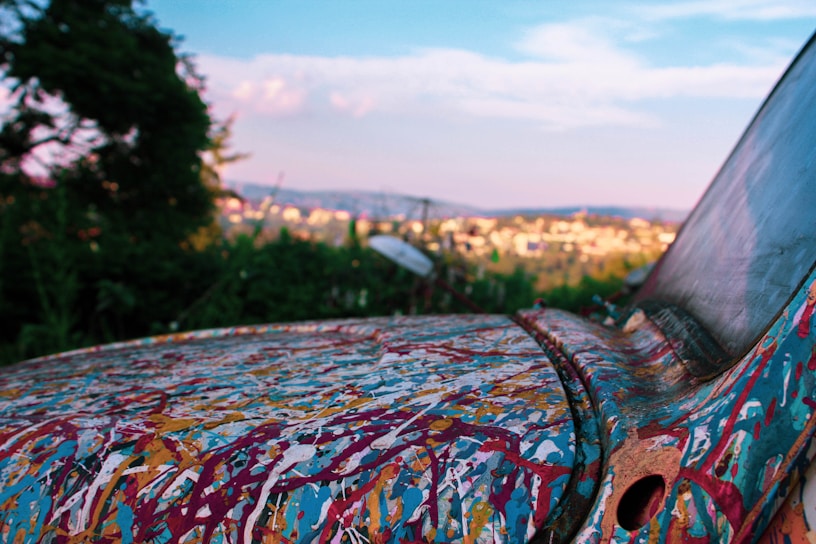 A paint-splattered, old car is parked in a natural setting, with a distant cityscape visible in the background. The vehicle's surface displays an abstract combination of colors against a backdrop of greenery and a blue sky.