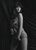 A monochrome image features a person posing with a cloth draped around their body. The background appears to be made of soft, flowing fabric, creating a sense of elegance and softness. The person exudes confidence, enhanced by the sophisticated drapery and lighting.