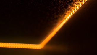 A diagonal line of warm, orange LED lights runs along a dark surface, creating a glowing effect that fades into the surrounding blackness. The lights appear to be in a strip form, subtly illuminating the area around them.