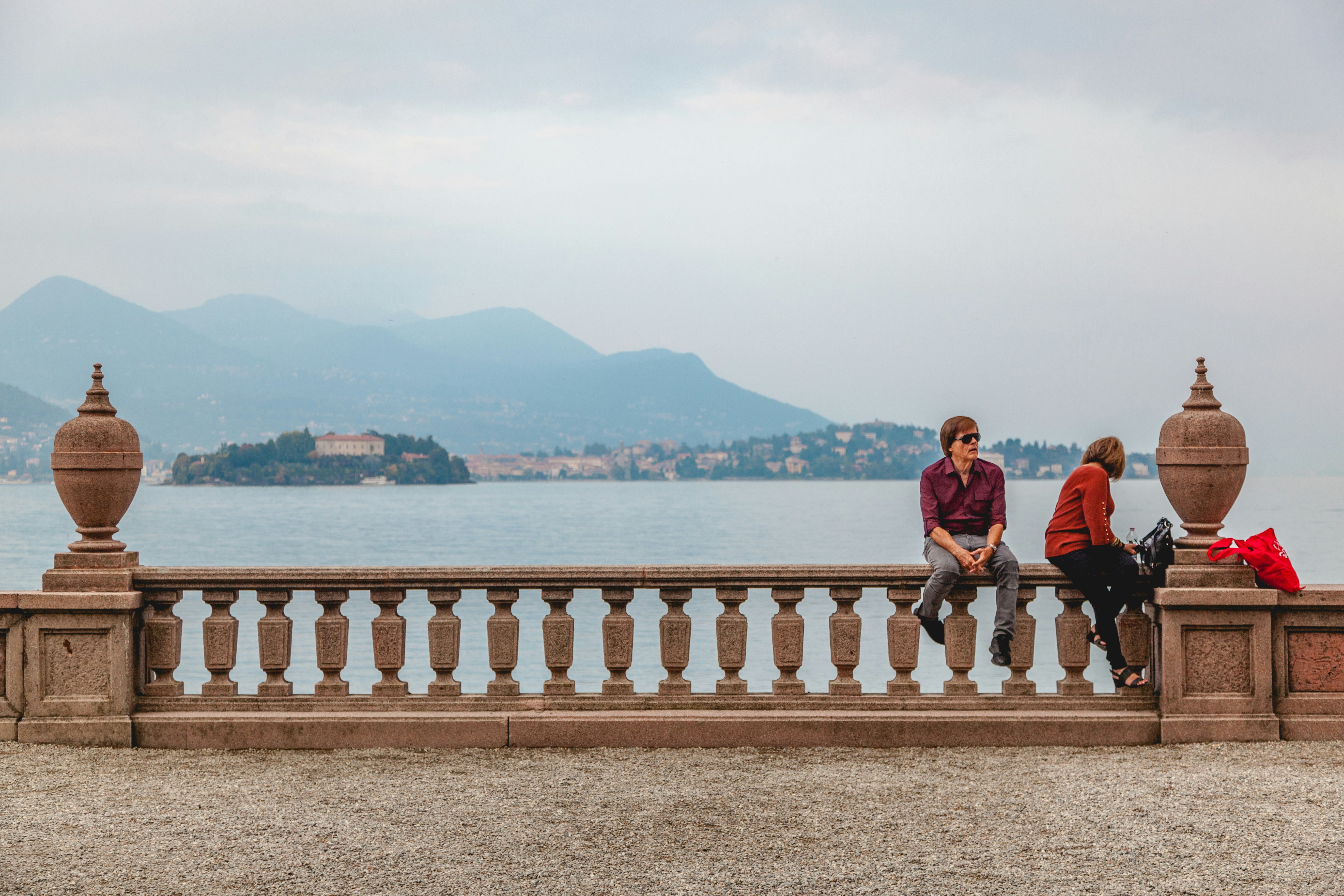 man and woman sitting on white wooden fence near body of water during daytime, Two people relaxing in front of "lago maggiore" on the "Isola Bella", beautiful island