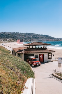 A lifeguard station situated by a scenic beach, featuring a red lifeguard vehicle parked in front. The station is topped with a tiled roof, and an American flag waves in the breeze nearby. The coastline curves into the distance against a backdrop of hills.