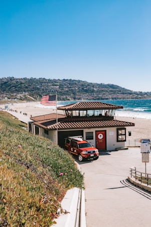 A lifeguard station situated by a scenic beach, featuring a red lifeguard vehicle parked in front. The station is topped with a tiled roof, and an American flag waves in the breeze nearby. The coastline curves into the distance against a backdrop of hills.
