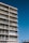 A tall apartment building with multiple floors and balconies. Each balcony is equipped with railings and some have furniture such as chairs and tables. The building is set against a clear blue sky, and palm trees can be seen in the distance.