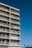A tall apartment building with multiple floors and balconies. Each balcony is equipped with railings and some have furniture such as chairs and tables. The building is set against a clear blue sky, and palm trees can be seen in the distance.