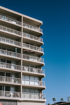 A tall apartment building with multiple floors and balconies. Each balcony is equipped with railings and some have furniture such as chairs and tables. The building is set against a clear blue sky, and palm trees can be seen in the distance.