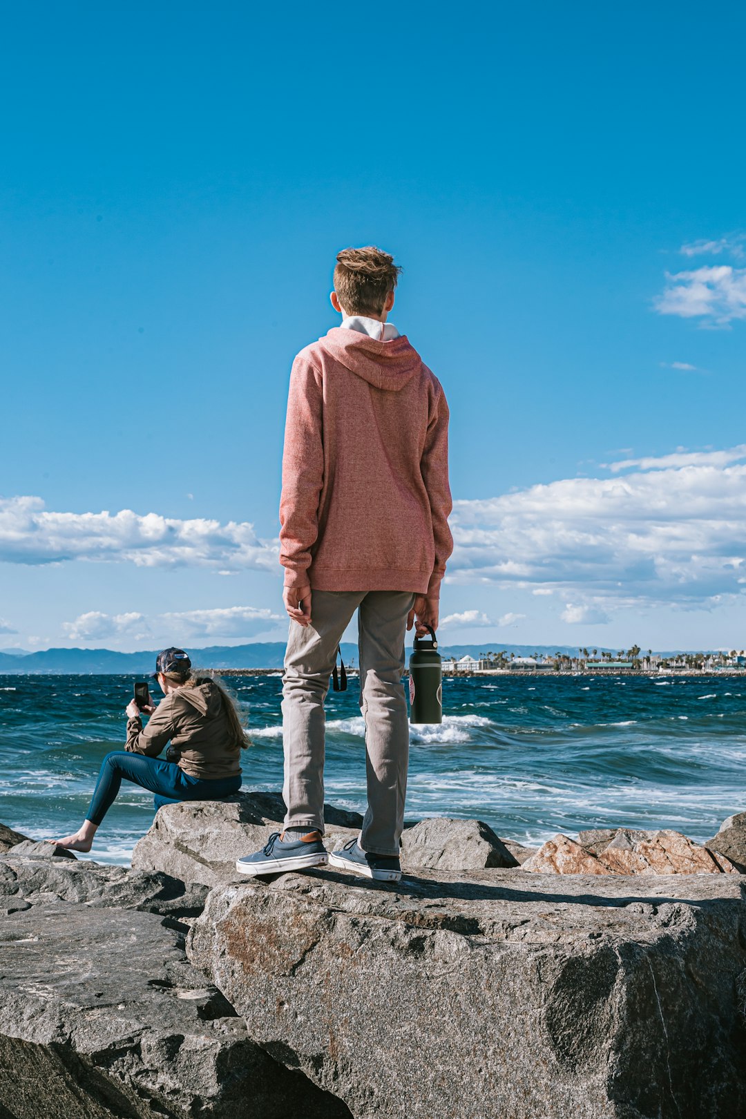 man in red sweater standing on rock near body of water during daytime