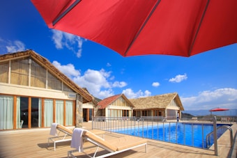 A bright poolside area with two modern sun loungers on a wooden deck, covered with white towels. The scene includes traditional thatched roofs of the buildings in the background, set against a vibrant blue sky dotted with fluffy white clouds. A large red umbrella provides shade at the forefront.