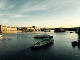 A scenic river view features two cruise boats sailing on a wide, tranquil river. The background includes a historic stone bridge and picturesque buildings lining the riverside, suggesting a European city setting. The sky is partially cloudy with warm sunlight casting a gentle glow over the scene.