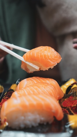 A close-up of several pieces of salmon sushi, with one piece being held by a pair of chopsticks above a decorative plate. The bright orange color of the salmon contrasts against the dark and intricate design of the plate.