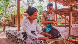 Two women are engaged in traditional craftwork outdoors, surrounded by tropical greenery and bamboo structures. The woman on the left is focused on weaving or preparing materials, while the woman on the right observes with a joyful expression. They are dressed in cultural attire, and a woven basket with colorful items is placed on the ground.