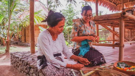Two women are engaged in traditional craftwork outdoors, surrounded by tropical greenery and bamboo structures. The woman on the left is focused on weaving or preparing materials, while the woman on the right observes with a joyful expression. They are dressed in cultural attire, and a woven basket with colorful items is placed on the ground.