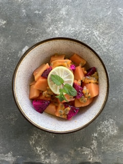 A ceramic bowl containing a fresh fruit salad with diced papaya, dragon fruit, and passion fruit seeds. A slice of lemon or lime with a couple of mint leaves is placed on top for garnish. The bowl is placed on a textured gray surface.