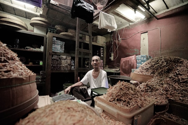 An older man sits surrounded by large quantities of dried goods, likely in a shop or storage area. The room is filled with shelves stocked with various packages and containers. The lighting is dim and there are exposed wires on the ceiling.