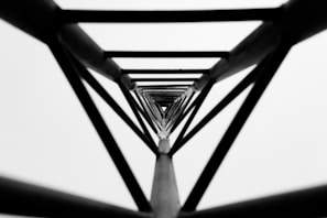 A symmetrical black and white perspective looking up through a series of concentric geometric frames, creating a tunnel-like effect. The frames form a repetitive, narrowing pattern that draws the viewer's eyes toward the center.
