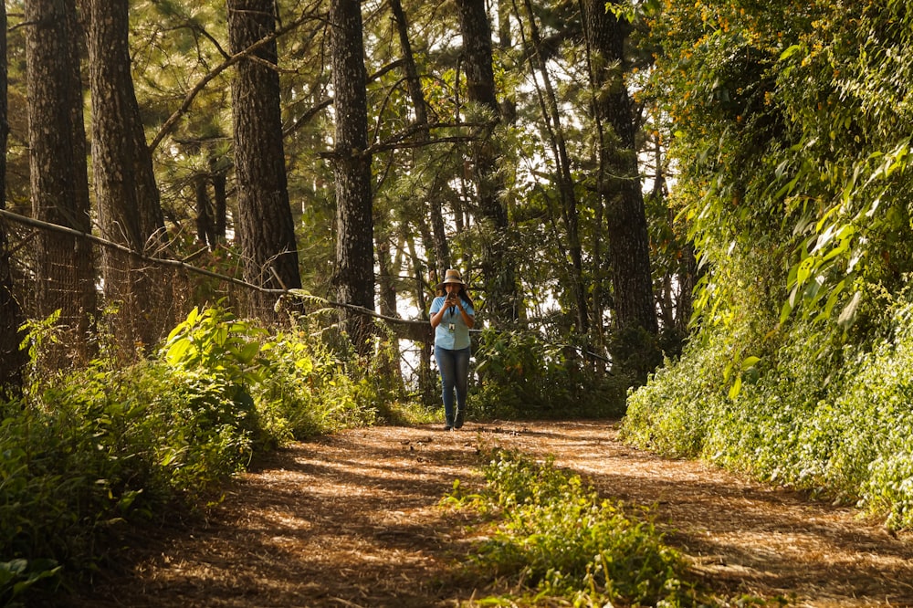 woman in blue jacket and blue denim jeans walking on pathway between green trees during daytime
