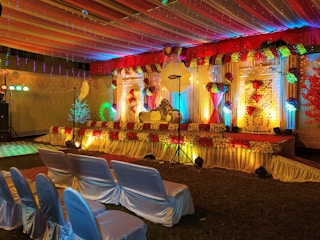 A beautifully decorated stage with vibrant floral arrangements and ornate backdrop. The stage is illuminated by colorful lights with red, yellow, and green hues. Chairs covered in white satin are arranged in rows facing the stage, and the ceiling is draped with multi-colored fabric, enhancing the festive atmosphere.
