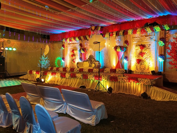 A beautifully decorated stage with vibrant floral arrangements and ornate backdrop. The stage is illuminated by colorful lights with red, yellow, and green hues. Chairs covered in white satin are arranged in rows facing the stage, and the ceiling is draped with multi-colored fabric, enhancing the festive atmosphere.