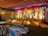 A beautifully decorated stage with vibrant floral arrangements and ornate backdrop. The stage is illuminated by colorful lights with red, yellow, and green hues. Chairs covered in white satin are arranged in rows facing the stage, and the ceiling is draped with multi-colored fabric, enhancing the festive atmosphere.